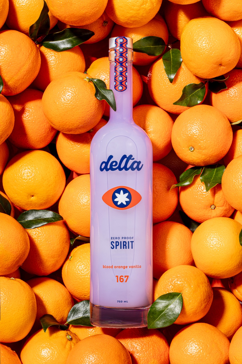 The Delta zero-proof Spirit bottle standing tall in a bed of fresh blood oranges with leaves still on, warm studio light, label clearly visible.